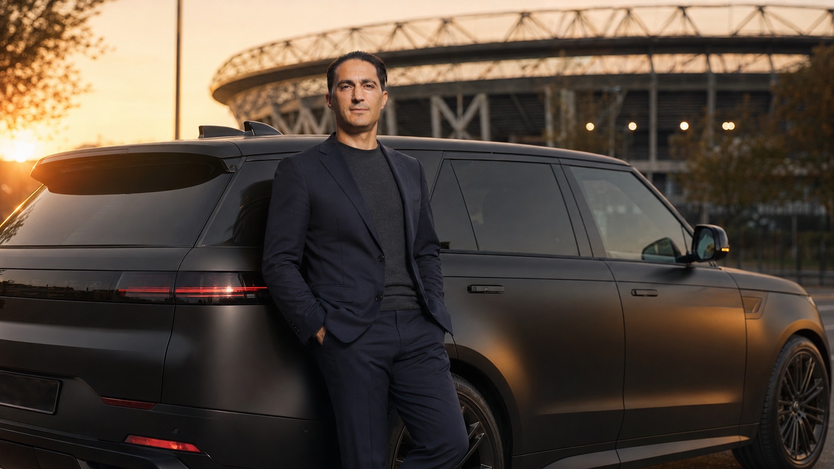 Serdar Gözübüyük in a dark suit, leaning against a matte-black Range Rover Sport outside a stadium at dusk.